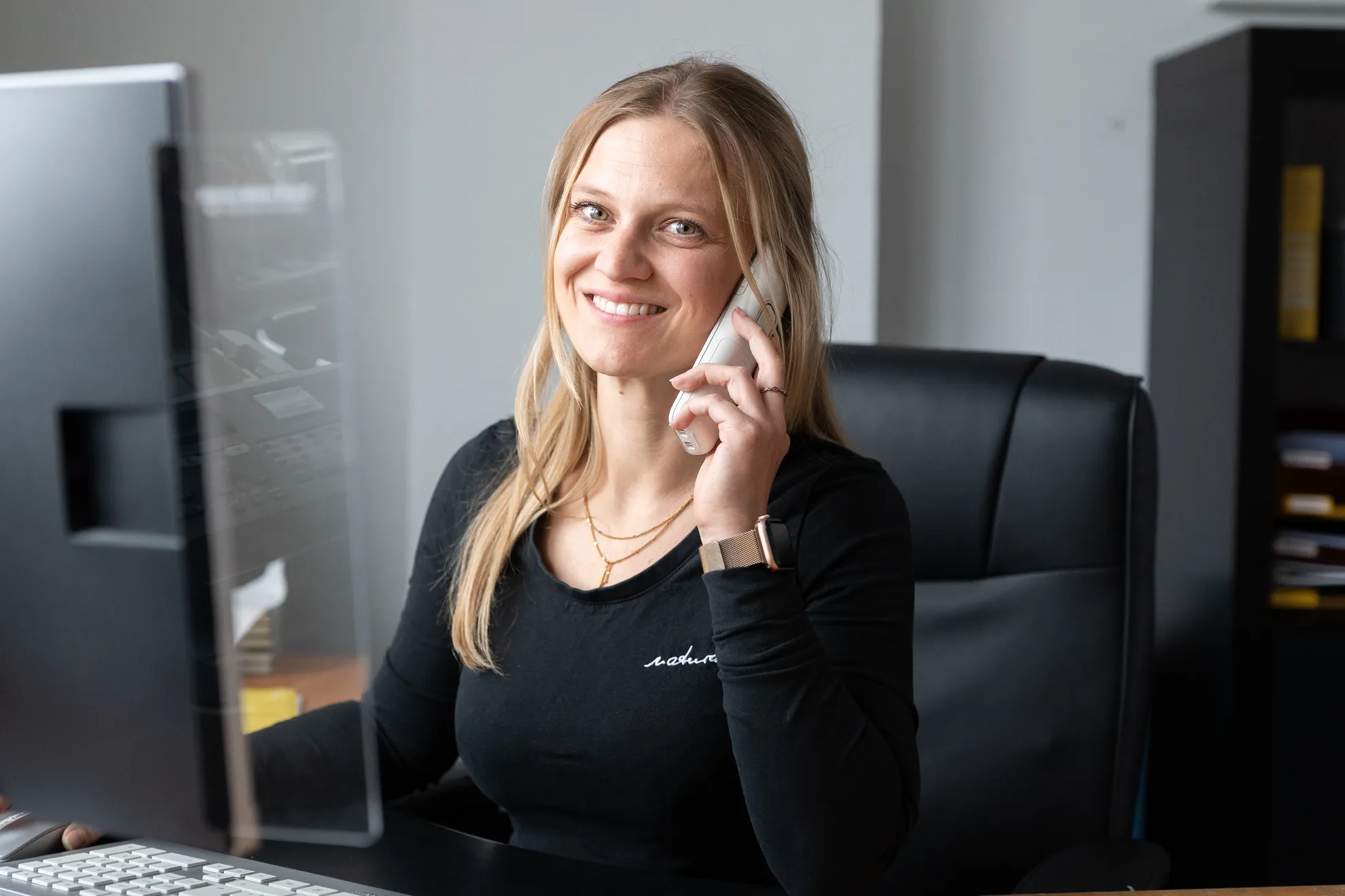 Eine Frau sitzt am Schreibtisch, lächelt in die Kamera und telefoniert