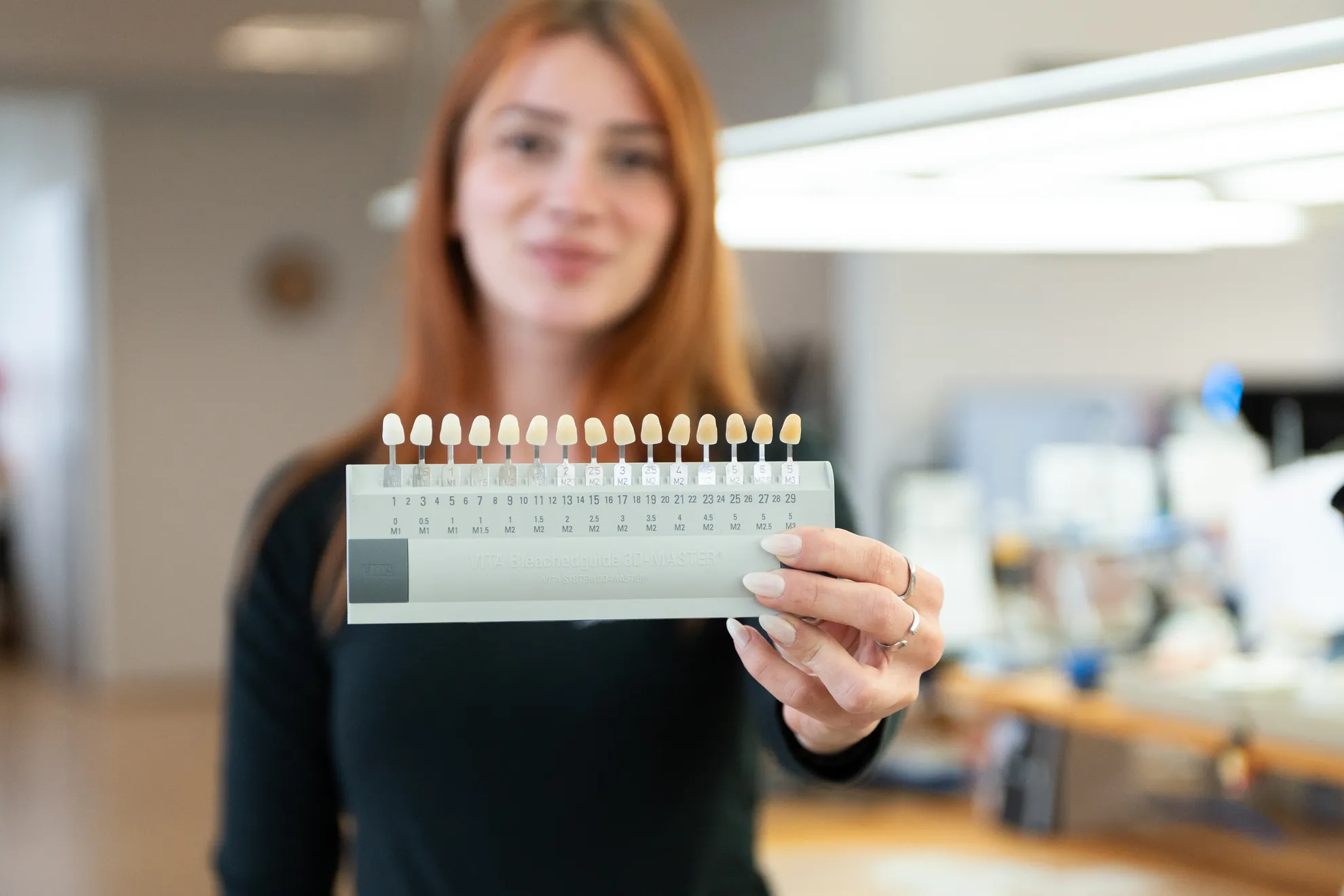 Eine Frau (unscharf im Hintergrund) hält eine Zahnfarbskala in die Kamera